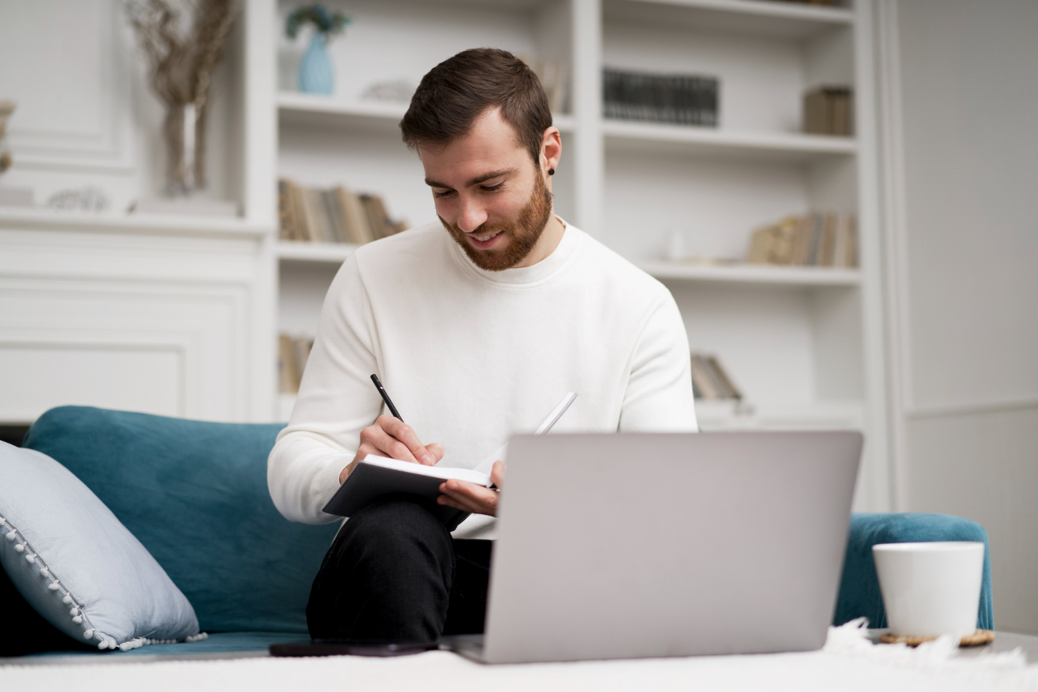 Hombre en sofá escribiendo en cuaderno con portátil. Suéter blanco, barba, estantería de fondo.