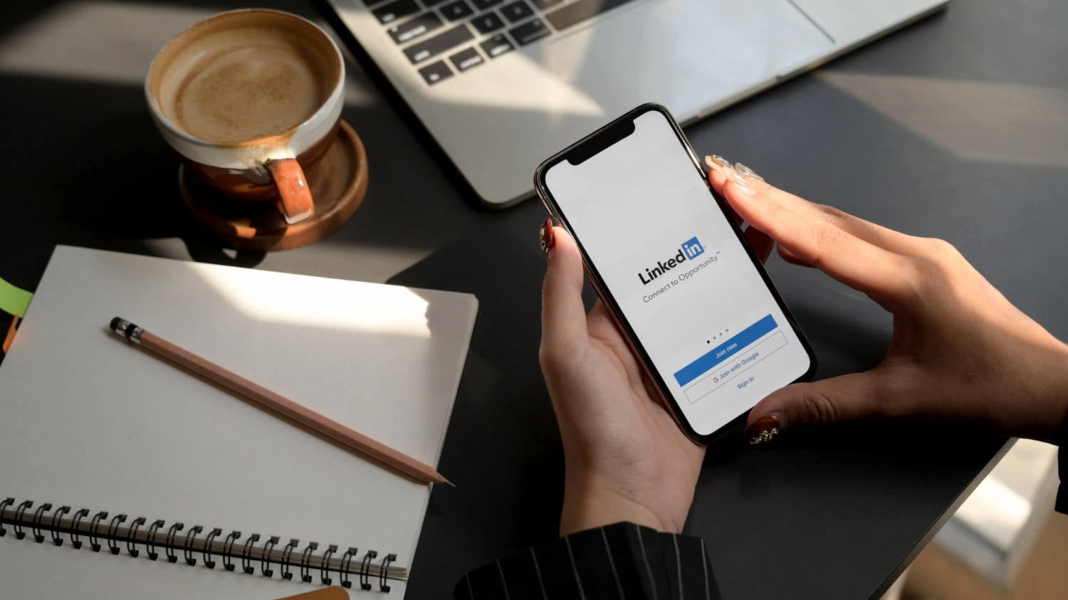 Escritorio con portátil, café, libreta y móvil mostrando la app de LinkedIn en manos de una persona.