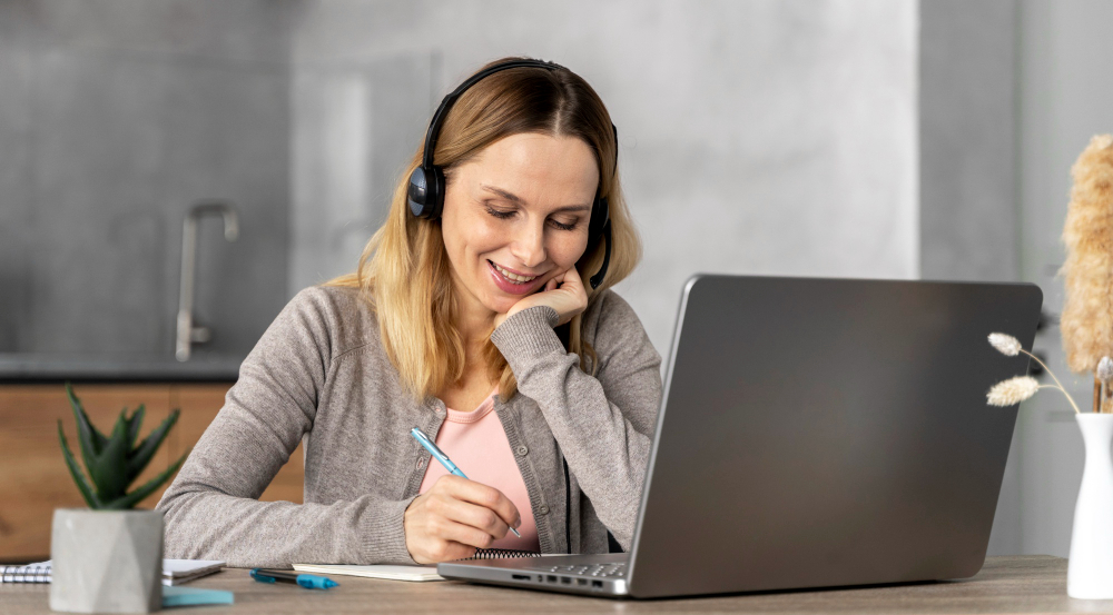 Mujer rubia con auriculares trabajando en portátil. Sonríe mientras escribe en un cuaderno.